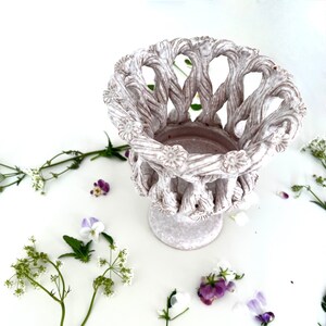 May include: White ceramic planter with a woven design and a wide, open top. The planter is surrounded by small white and purple flowers.