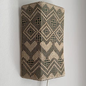 Può includere: Lampada da parete rettangolare con un motivo geometrico beige e verde scuro. La lampada presenta un design traforato e tessuto, e un cavo bianco con un interruttore on/off. È montata su una parete bianca.