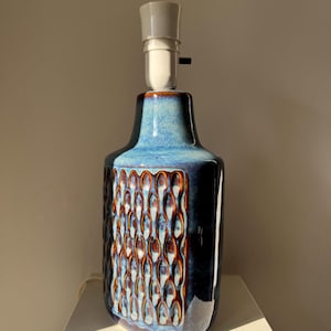 May include: A ceramic table lamp with a blue and brown glaze. The lamp has a textured pattern on the front and a white lamp holder. The lamp is on a white surface.
