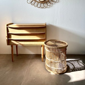 May include: A wooden chest of drawers with three drawers and a woven wicker side table. The dresser and table are both light brown in colour. A woven rattan eye-shaped mirror hangs on the wall behind the furniture.