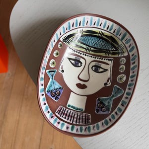 May include: An oval ceramic dish with a brown background featuring a stylized face illustration in black and white. The design includes two blue fish motifs, a patterned hat, and a decorative border with alternating blue and pink dash markings.