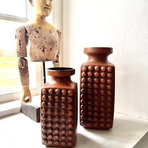 Può includere: Due vasi in ceramica marrone con un motivo strutturato e puntinato. I vasi sono rettangolari con un collo stretto e un interno scuro. Sullo sfondo c'è un manichino di legno vintage.