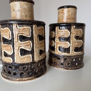 May include: Two ceramic vases with a cylindrical shape. The vases have a textured, beige, and black pattern with a perforated base. The top of the vases are beige with a dark brown rim.