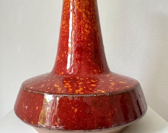 Może przedstawiać: Ceramiczna lampa stołowa z czerwoną i pomarańczową plamistą glazurą. Lampa ma szeroką, płaską podstawę, rozszerzany korpus i wąską szyję prowadzącą do białego uchwytu lampy. Lampa jest w stylu vintage.