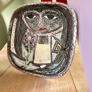 Puede incluir: Cuenco de cerámica pintado a mano con un diseño de rostro estilizado. El cuenco presenta un fondo oscuro con una figura central, una flor y patrones geométricos en varios colores. El borde tiene un borde decorativo.