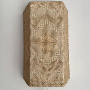 May include: A rectangular wall lamp featuring a beige crochet lace cover with a diamond-shaped pattern. The light fixture has a structured frame underneath the fabric, with a white power cord extending from the bottom edge of the lamp.