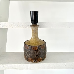 Könnte beinhalten: Eine Keramik-Tischlampe mit einem braunen und beigefarbenen, strukturierten Sockel und einem schwarzen Lampenschirm. Die Lampe steht auf einem weißen Regal.