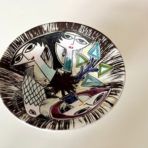 May include: A white ceramic bowl with a black and white striped border. The bowl features a colourful abstract design with stylised faces and geometric shapes.