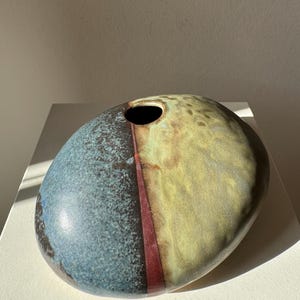 May include: A rounded, flattened ceramic vase with a smooth, glazed finish. The surface features a split color design with a textured blue-grey section on the left and a mottled olive-green section on the right, separated by a thin, dark red vertical line.