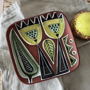 May include: A square, ceramic plate with a reddish-brown background. The plate features a stylized floral design in yellow, black, and green. A small, round dessert with a yellow glaze and nut crust sits nearby.