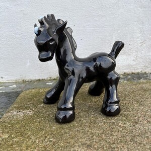 May include: A glossy black ceramic horse figurine with a stylized design. The horse has a rounded body, short legs, and a small tail. It has light blue eyes and a white patch on its head. The figurine is standing on a stone surface.