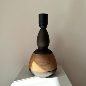 Könnte beinhalten: Ein handgefertigter Lampensockel mit schwarzem Oberteil und dunklem, strukturiertem Körper. Der Sockel hat ein Band aus gewebtem, hellbraunem Seil um die Mitte. Der untere Teil ist dunkler, metallisch gefärbt. Die Lampe ist für eine Glühbirne konzipiert.