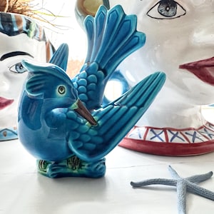 Op de afbeelding: Een blauw keramiek vogeltje met een lange snavel en een gespreide staart. Het vogeltje zit op een wit oppervlak met een wit keramiek hoofd en een zeester.