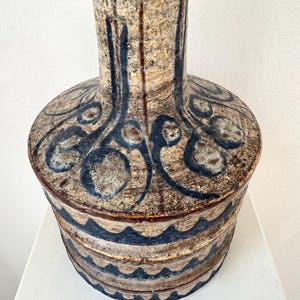 May include: A handcrafted ceramic vase with a textured surface. The vase features a cylindrical base with a series of blue wave-like patterns. The upper portion has a wider, rounded section with blue abstract designs. The vase is primarily beige and brown.