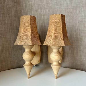 Könnte beinhalten: Ein Paar Wandleuchten aus hellem Holz. Jede Lampe verfügt über einen ausgestellten, sechseckigen Schirm mit markanten kreisförmigen Holzmaserungsmustern und einen gedrechselten, spindelförmigen Sockel mit einem spitz zulaufenden, konischen Ende.