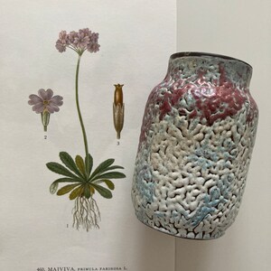 May include: A white ceramic vase with a blue and pink mottled glaze. The vase has a textured surface and a rounded shape. The vase is sitting on a white surface.