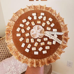 May include: A round, brown sweet potato pie with a decorative crust and a dusting of cinnamon. The pie is topped with white marshmallows and a cream-colored rosette. A small tag reads "Sweet potato".