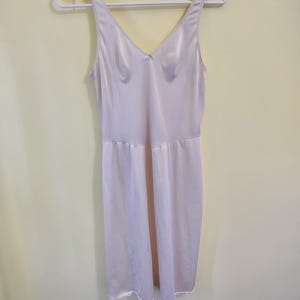May include: A pale pink slip dress with a V-neck and spaghetti straps hangs on a white hanger. The dress has a fitted bodice, a small bow detail, and a flowing skirt with a lace trim at the hem. The fabric appears to be satin.