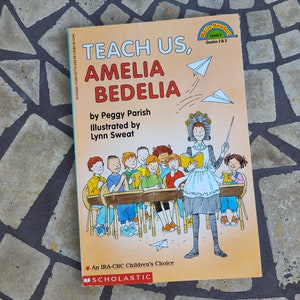 Puede incluir: Un libro infantil titulado "Teach Us, Amelia Bedelia" de Peggy Parish, ilustrado por Lynn Sweat. La portada presenta una ilustración de Amelia Bedelia en un aula con estudiantes. El libro es un libro Hello Reader de nivel 4 para los grados 2 y 3.