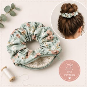 May include: A sage green fabric scrunchie with a floral pattern featuring pink and white flowers and green leaves. The scrunchie includes a hidden zipper pocket and is shown worn in a hair bun. A spool of white thread is visible nearby.