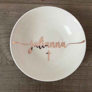 May include: A white ceramic bowl with the name "Julianna" in rose gold script and a rose gold cross. The bowl has a simple, elegant design, suitable for serving or display.