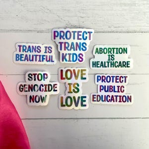 May include: A collection of holographic stickers with various social justice messages. The stickers feature phrases like "Protect Trans Kids," "Abortion is Healthcare," and "Love is Love" in colorful, bold fonts. The stickers are on a white wooden surface.