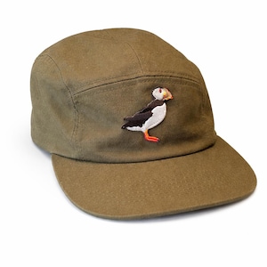Puede incluir: Gorra de cinco paneles verde oliva con visera plana. Un parche bordado de frailecillo está en la parte delantera. El frailecillo tiene pecho blanco, espalda negra y patas y pico naranjas. La gorra está hecha de tela duradera.