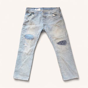 Może przedstawiać: Jasnoniebieskie dżinsy z naszywkami na kolanach. Dżinsy mają zapięcie na guziki i klasyczny krój z pięcioma kieszeniami. Dżins jest zniszczony z pewnym wyblaknięciem i małymi rozdarciami.