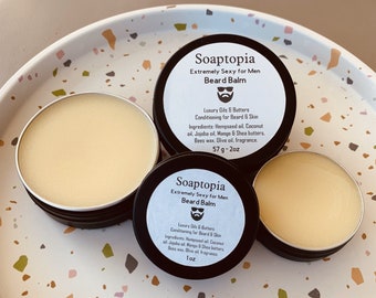Può includere: Due piccoli contenitori rotondi di balsamo per barba, entrambi etichettati "Soaptopia Extremely Sexy for Men Beard Balm". I contenitori sono su una superficie macchiata di bianco e marrone. Gli ingredienti e le dimensioni sono elencati sulle etichette.