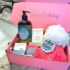 May include: Pink gift box with a Happy Birthday! message on the lid. The box contains a variety of bath and body products, including a bottle of liquid soap, a bar of soap, a bath puff, and a small decorative sign that says "enjoy today".