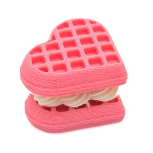 Op de afbeelding: Een roze hartvormige wafel sandwich met witte crèmevulling. De wafel heeft een rasterpatroon en is gemaakt van een textuurrijk materiaal. Het dessert staat tegen een witte achtergrond.