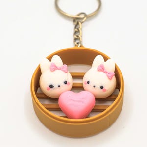 May include: A keychain featuring two white bunny-shaped steamed buns with pink bows, a pink heart-shaped bun, and a gold-colored bamboo steamer.