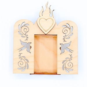 May include: A wooden frame with a heart cutout and a flame design at the top. The frame has a cutout of two birds and floral designs on each side. The frame is made of light wood and has a natural finish.