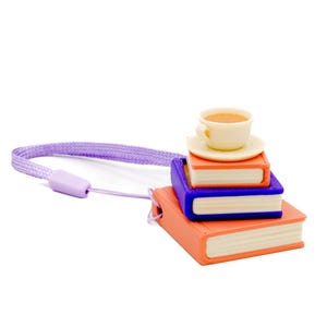 Pode incluir: Uma pilha em miniatura de livros laranja e azuis encimada por uma pequena chávena e pires. Uma fita roxa está presa aos livros. A chávena contém um líquido castanho claro.
