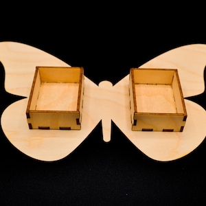 May include: Two small, rectangular wooden boxes sit on a butterfly-shaped wooden base. The boxes are light brown with darker brown accents. The butterfly base is also light brown. The background is black.