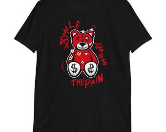 May include: Black t-shirt with a red teddy bear graphic. The teddy bear has a stitched-up face and dollar signs on its feet. The text "Smile Through The Pain" is written in red around the bear.