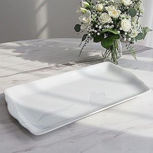 Puede incluir: Una fuente rectangular blanca con un sutil diseño floral. La fuente tiene bordes suavemente festoneados y está hecha de un material brillante. Un jarrón con flores blancas se encuentra en el fondo.