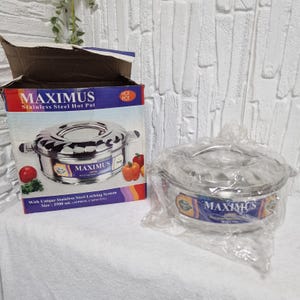Puede incluir: Se muestra una olla de acero inoxidable de la marca "MAXIMUS". La olla es redonda con un asa y está envuelta en plástico transparente. También se ve la caja del embalaje, con el nombre del producto y una imagen de la olla.