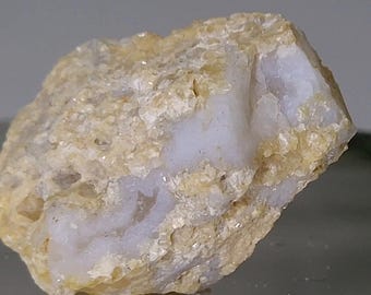 May include: A raw, unpolished mineral specimen featuring a rough, textured exterior in shades of pale yellow and beige. The stone displays crystalline, translucent white inclusions embedded within the porous, sandy-colored rock formation.