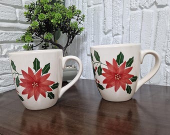 May include: Two off-white ceramic mugs with large red poinsettia flower designs and green leaves. The mugs have curved handles and are displayed on a dark wooden surface. The floral design is hand-painted, creating a festive holiday theme.