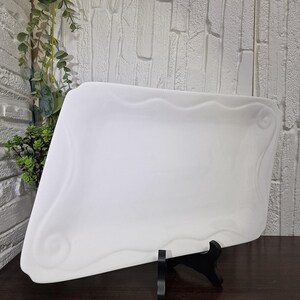 Puede incluir: Una fuente rectangular de cer&aacute;mica blanca con un borde decorativo ondulado. La fuente est&aacute; sostenida por un soporte negro, mostrando su dise&ntilde;o. Ideal para exhibir alimentos.