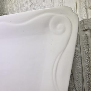 Puede incluir: Primer plano de un objeto rectangular blanco con un dise&ntilde;o de remolino decorativo curvo en la esquina. El objeto tiene un acabado liso y brillante. El fondo es una pared de ladrillo gris claro.