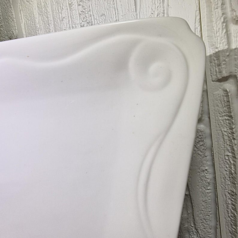 Puede incluir: Primer plano de un objeto rectangular blanco con un dise&ntilde;o de remolino decorativo curvo en la esquina. El objeto tiene un acabado liso y brillante. El fondo es una pared de ladrillo gris claro.
