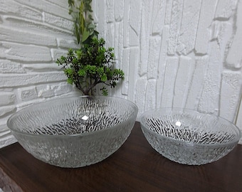 May include: Two clear glass bowls with a textured, organic pattern. The bowls are different sizes and sit on a dark brown surface. The background features a white brick wall and a small green plant.