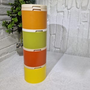 Könnte beinhalten: Ein Stapel von vier bunten, zylindrischen Behältern. Die Behälter sind in den Farbtönen Orange, Grün, Rotorange und Gelb gehalten und haben cremefarbene Deckel. Die Behälter sind übereinander gestapelt.