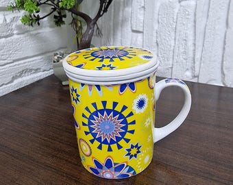 May include: Yellow ceramic mug with a matching lid, adorned with a vibrant blue and white floral pattern. The mug has a white handle and a white rim on the lid. The design is repeated on the lid and the body of the mug.