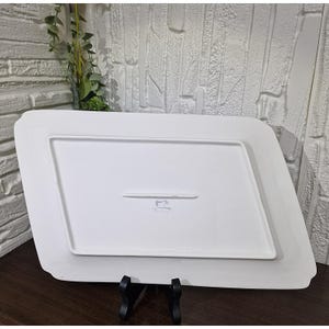 Puede incluir: Una fuente rectangular blanca con un dise&ntilde;o ligeramente angulado. La fuente est&aacute; sostenida por un soporte negro, mostrando su parte inferior con un sutil emblema en relieve. El fondo presenta una pared de ladrillos blancos texturizados.