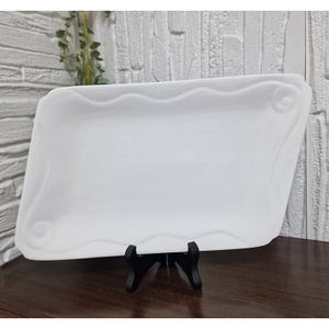 Può includere: Un vassoio rettangolare bianco con un bordo ondulato e decorazioni a volute agli angoli. Il vassoio è esposto su un supporto nero, che ne mette in risalto la forma elegante e l'estetica pulita. Lo sfondo presenta un muro strutturato.