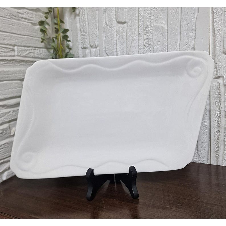 Puede incluir: Una fuente rectangular blanca con un dise&ntilde;o de borde ondulado y remolinos decorativos en las esquinas. La fuente se exhibe sobre un soporte negro, mostrando su elegante forma y est&eacute;tica limpia. El fondo presenta una pared texturizada.