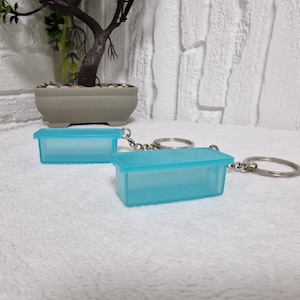 May include: Two turquoise rectangular keyrings. Each keyring features a small, box-shaped container with a textured lid. The keyrings are attached to silver key rings and chains. The background is a white, textured surface.